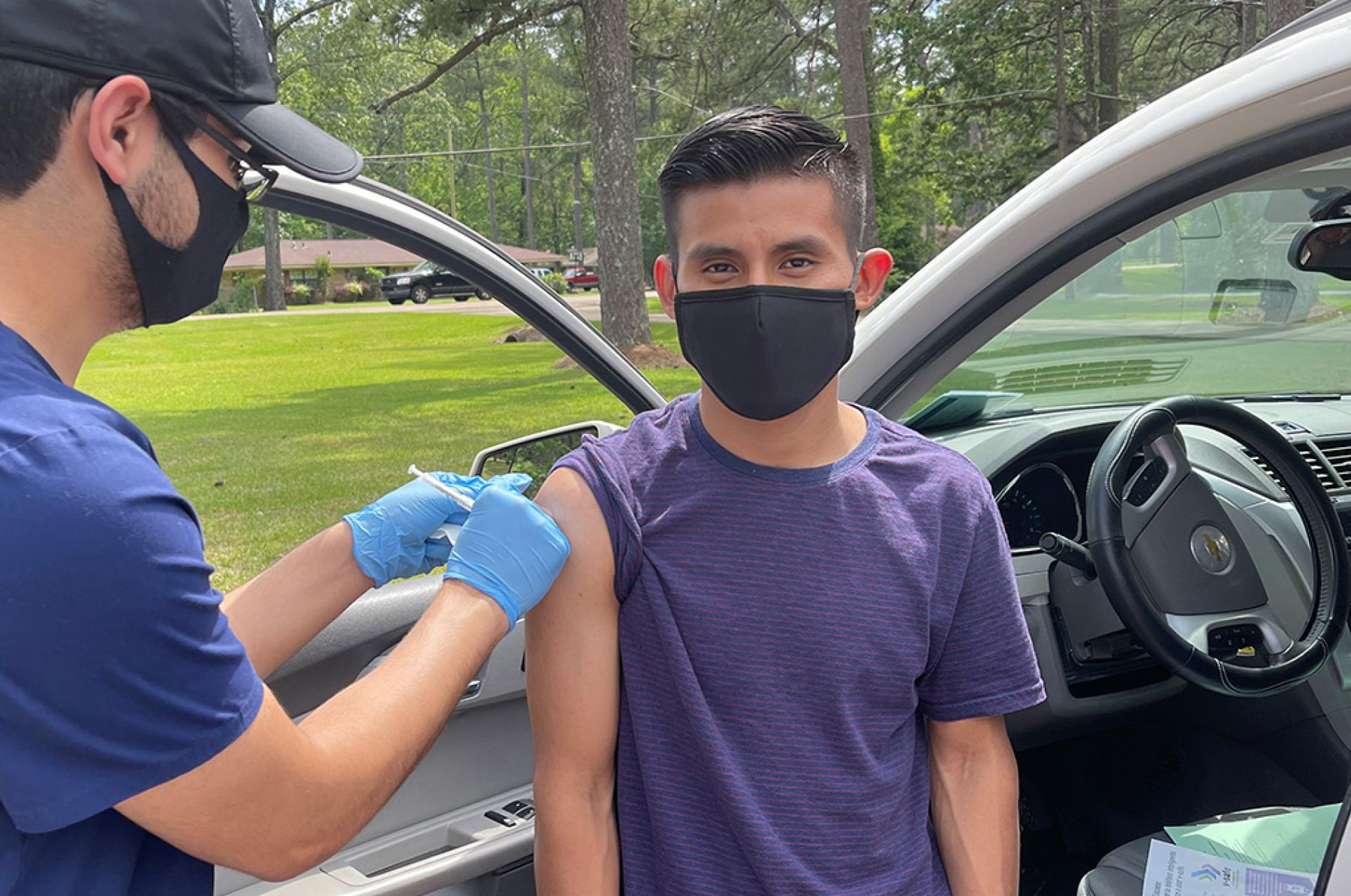 Masked teenage boy stands by a car while receiving a vaccine in his arm from a masked provider.