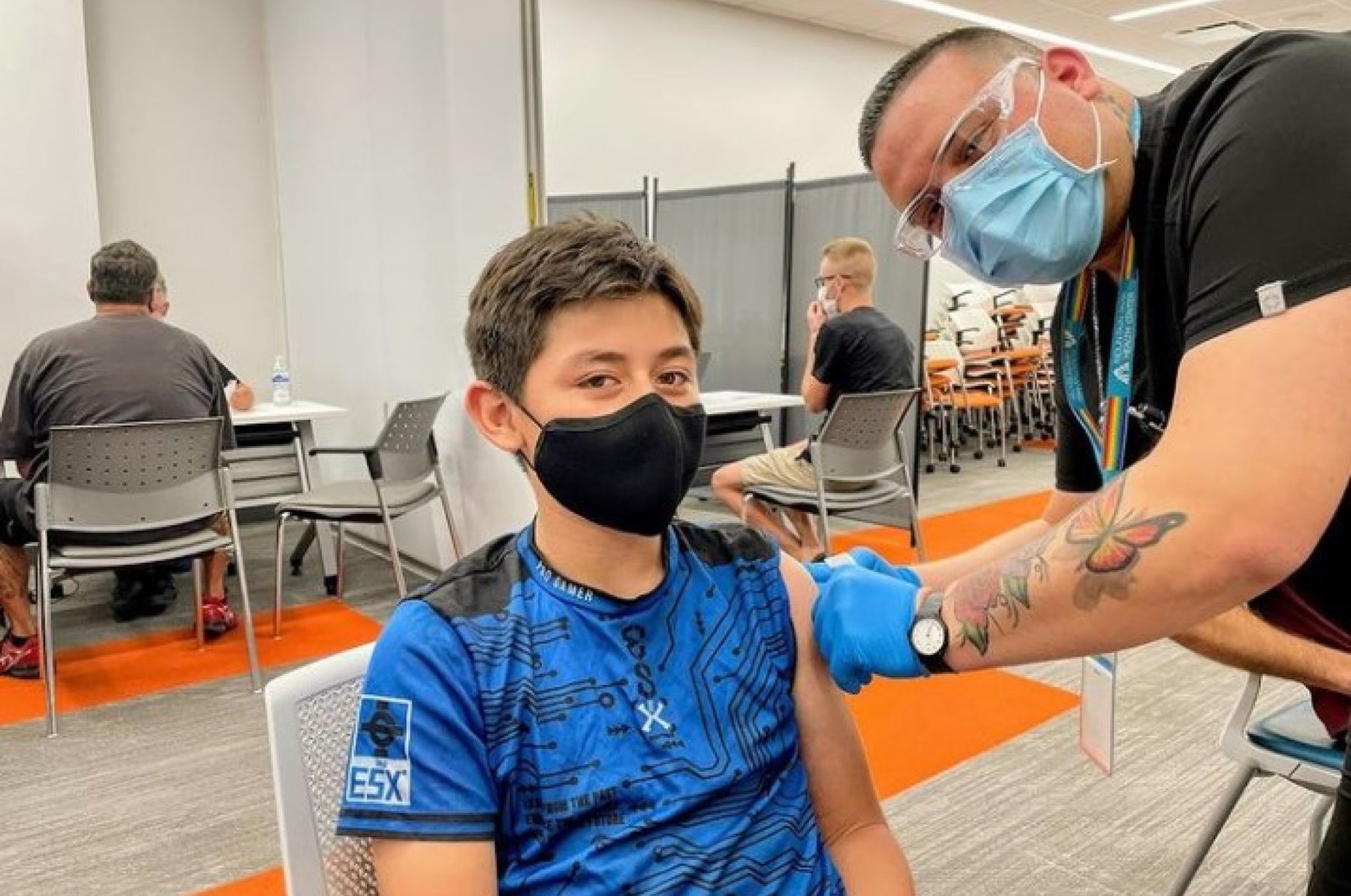 Masked tween boy sits in a medical center and receives a vaccine from a masked provider with tattoos. 