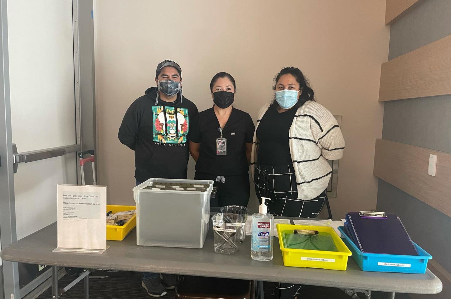 Three masked people with ID badges stand behind a check-in table with files, pens, clipboards, and hand sanitizer.