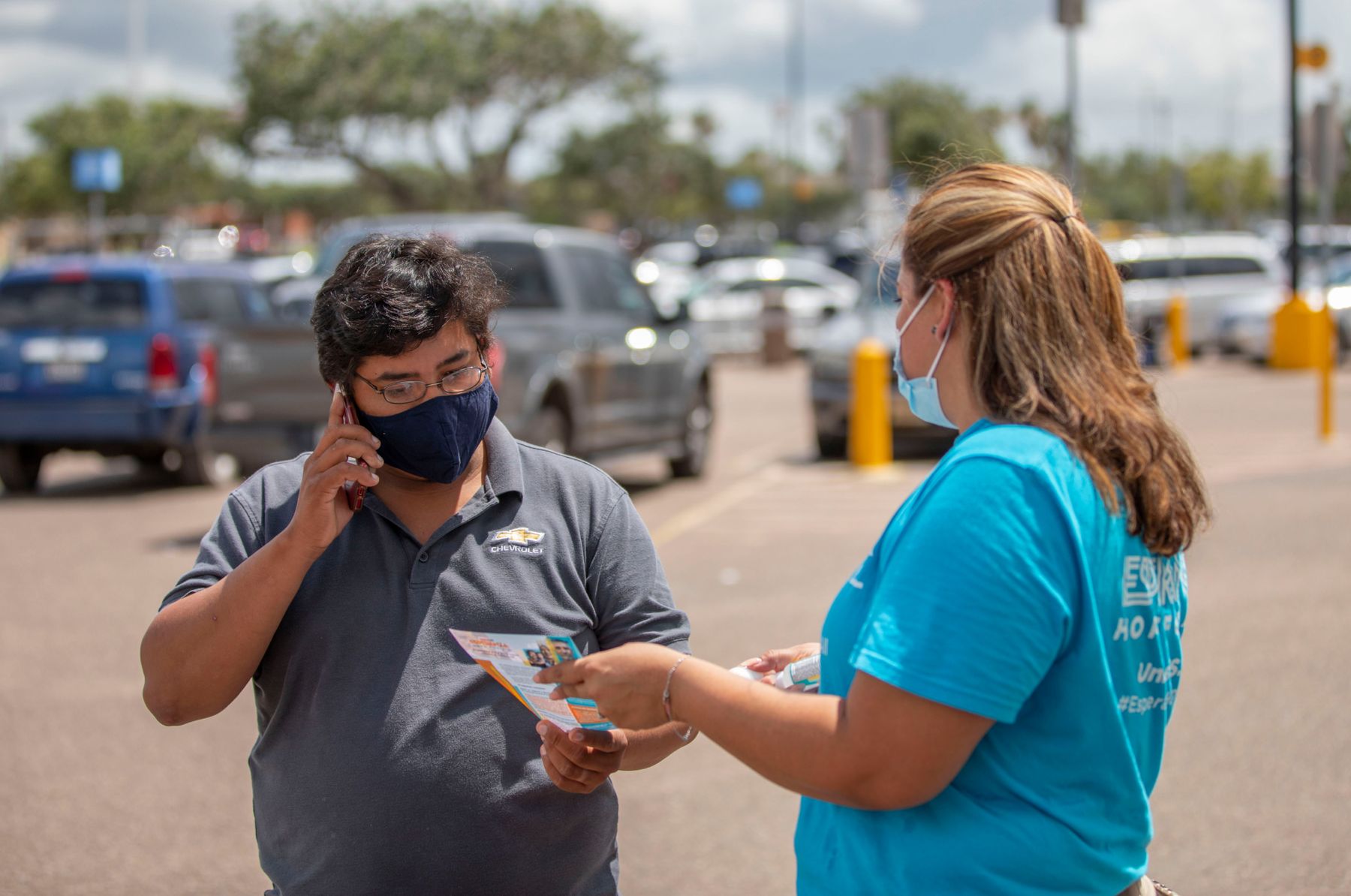 Masked woman working at an Esperanza event shows a Covid vaccine pamphlet to a masked man talking on a phone.