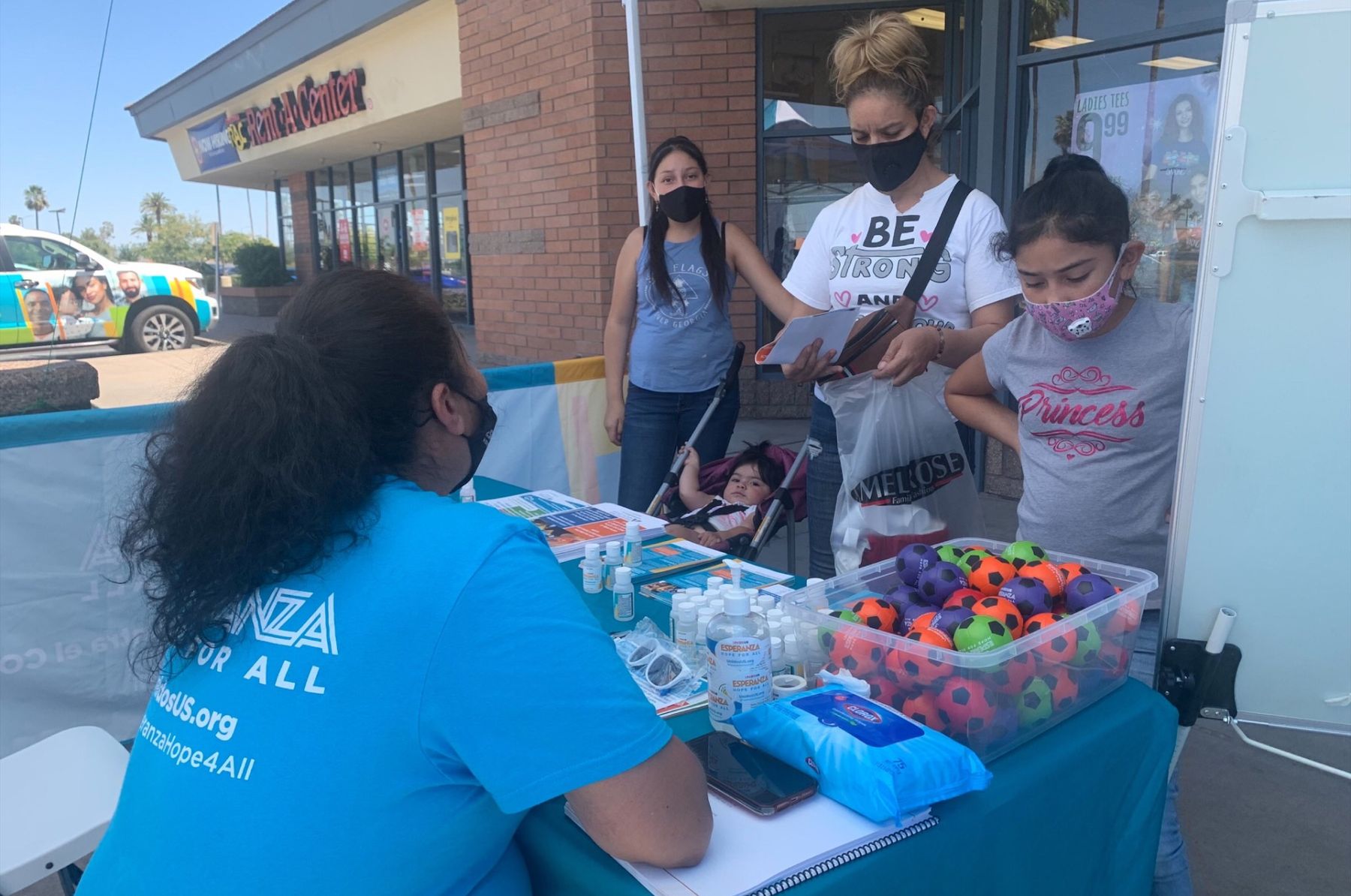 A mom and three daughters speaks with a woman working at an Esperanza table that has hand sanitizer and toy balls.