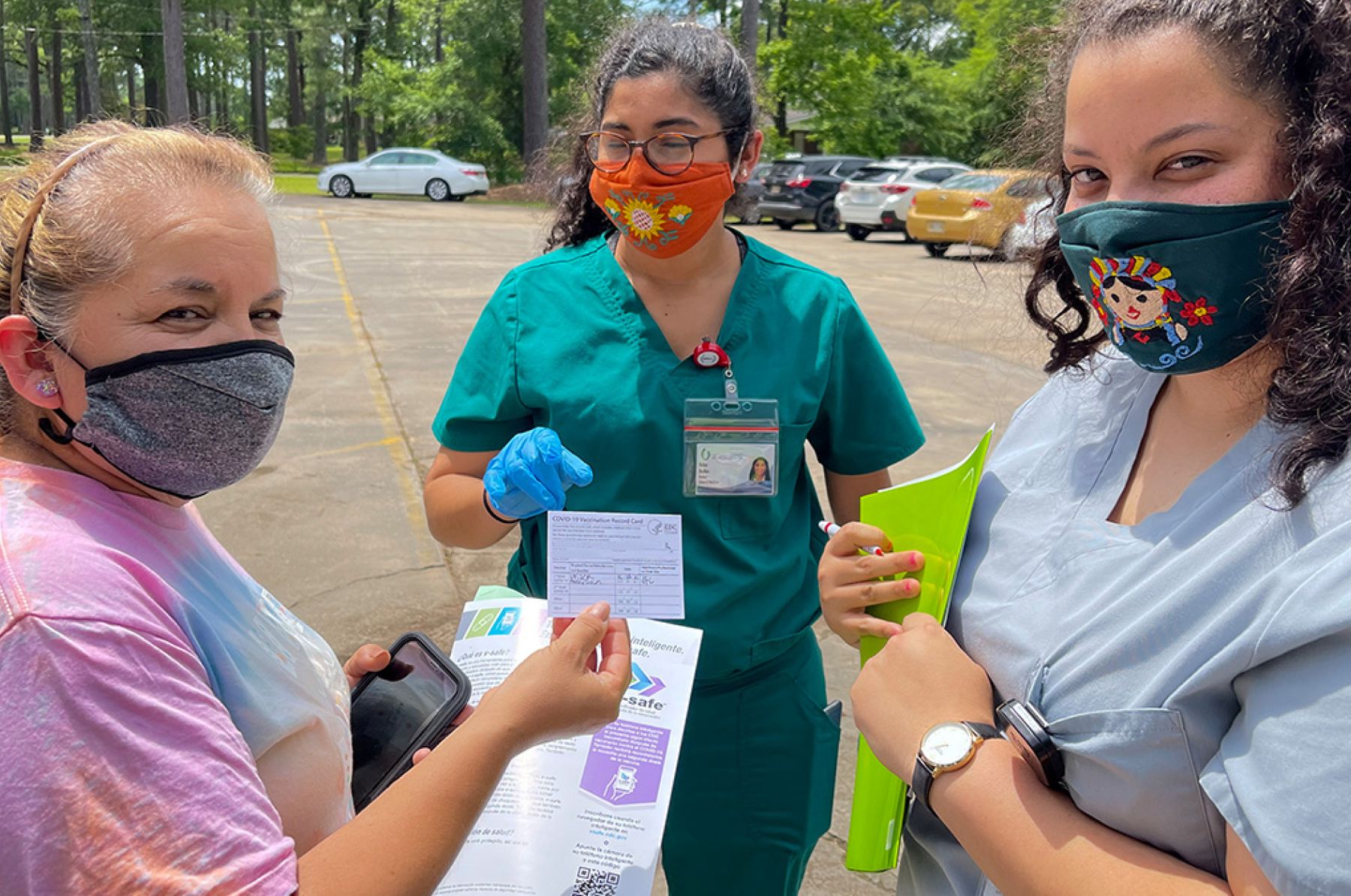 Two masked nurses in scrubs stand with a masked woman holding up her Covid vaccine card in a parking lot.