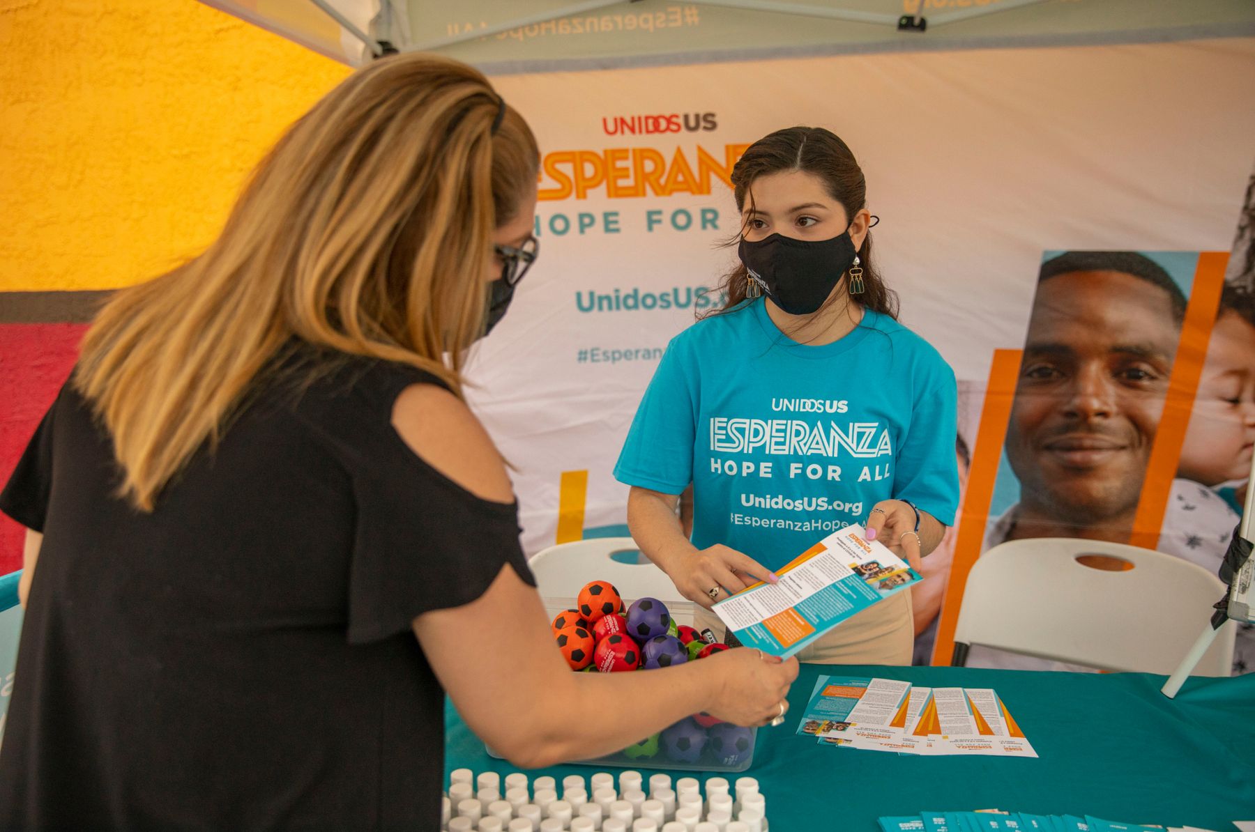 Masked woman working at the Esperanza tent shows another masked woman a pamphlet about the Covid vaccine.