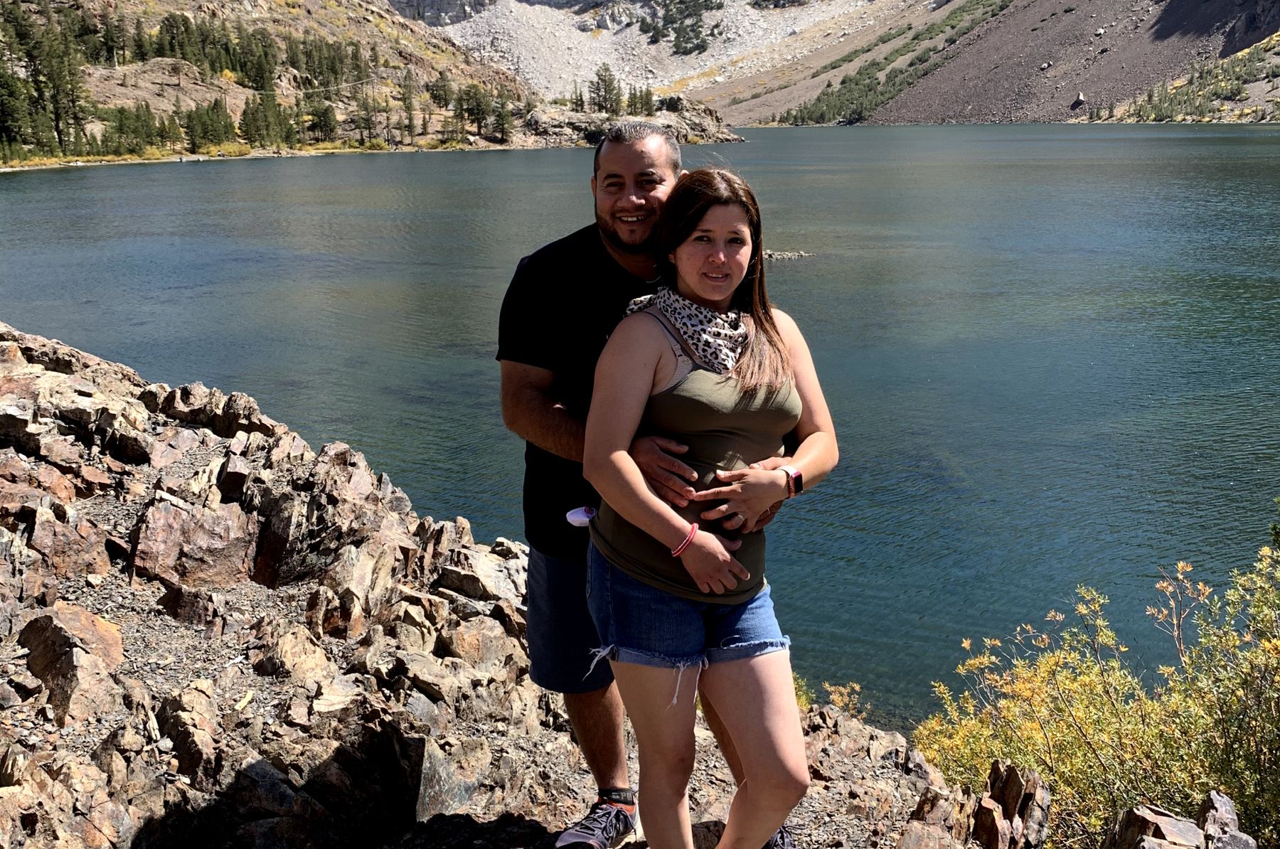 Husband and pregnant wife stand together with hands on her stomach in front of a lake.