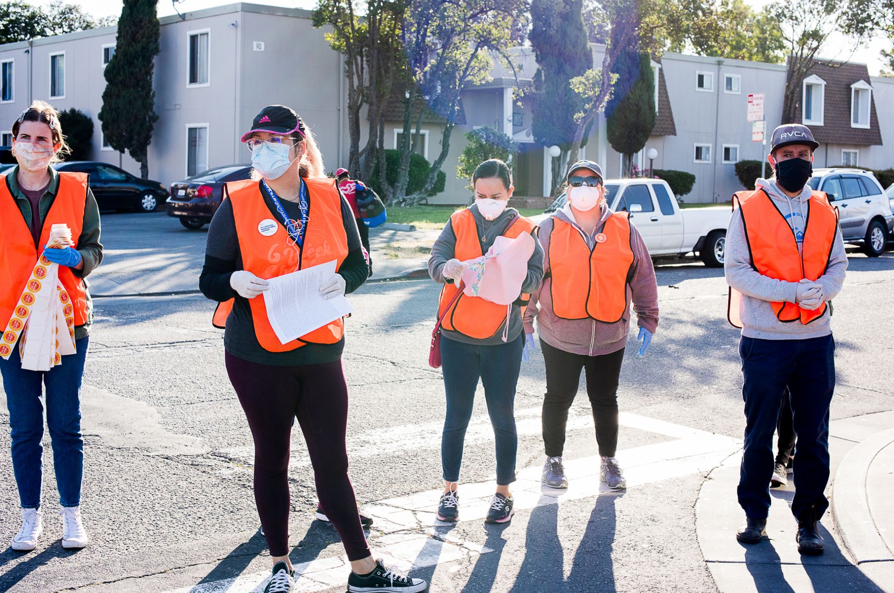 A group of people wearing orange safety vests, masks, and gloves stand on a city street on a sunny day.