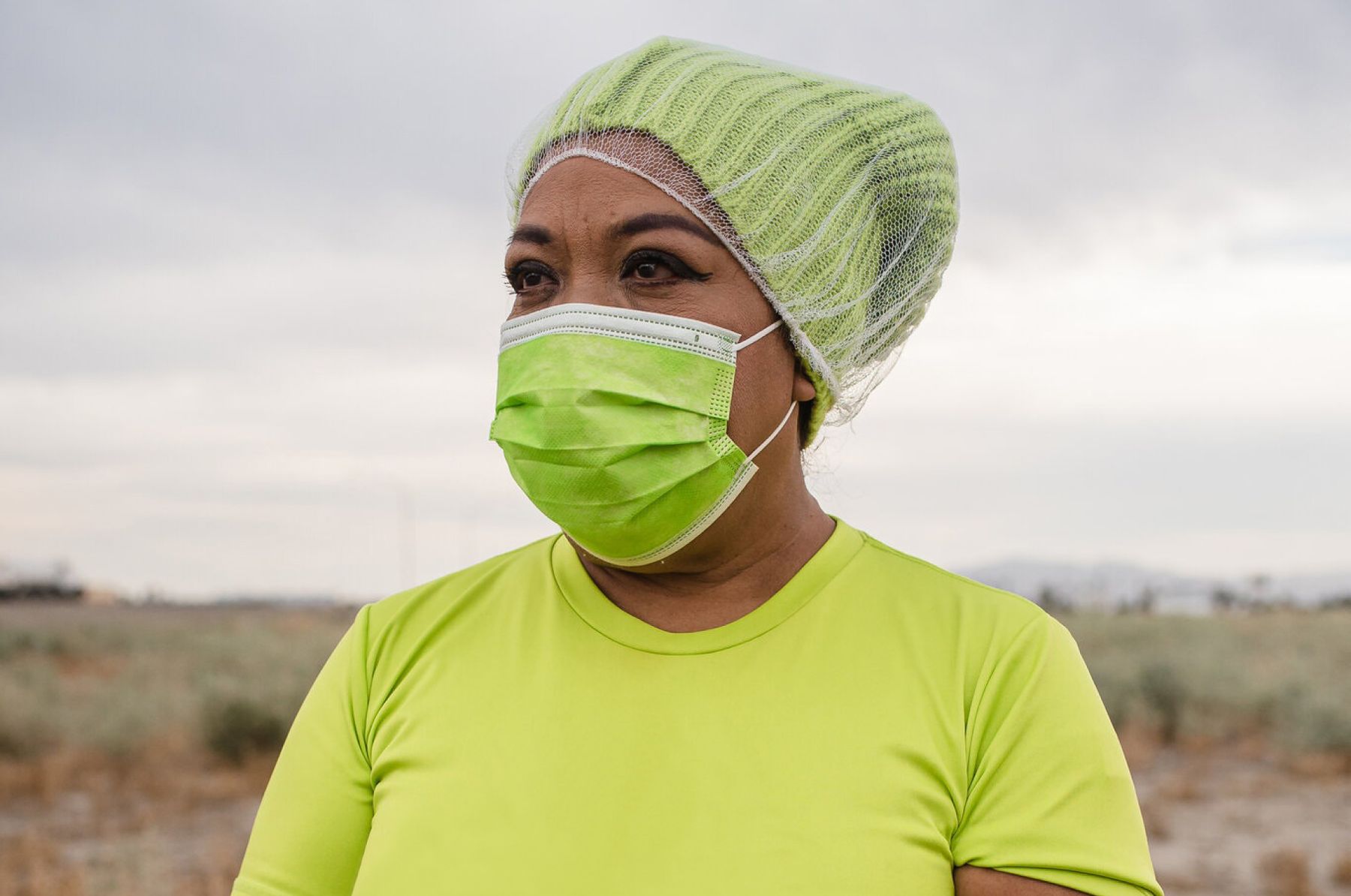 Woman wearing a yellow mask and a knit hat with a hairnet stands in front of a farm field and looks in the distance.