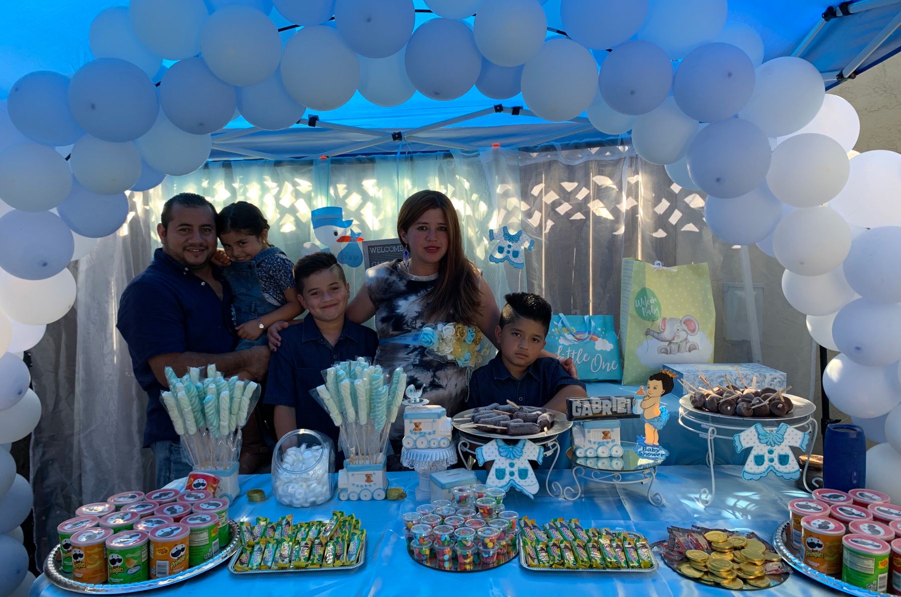 Smiling family with three boys celebrating at a baby shower with a blue baloon arch and a table full of candy and snacks. 