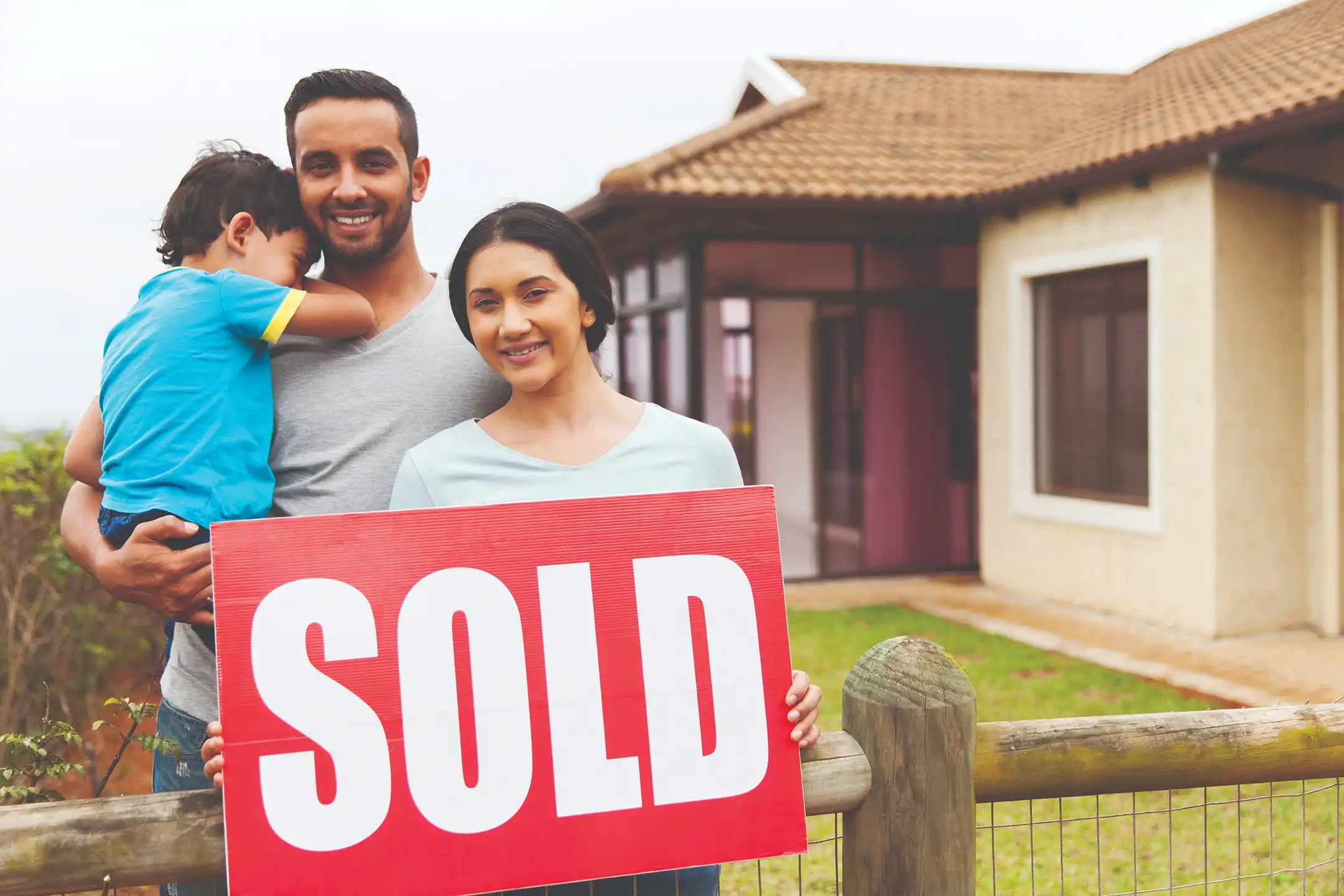 A family of three holding a "sold" sign outside their newly purchased home.